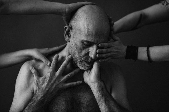 Dynamisches Porträt eines älteren Mannes, dessen Gesicht von verschiedenen Händen umgeben und berührt wird, was die vielen Seiten menschlich